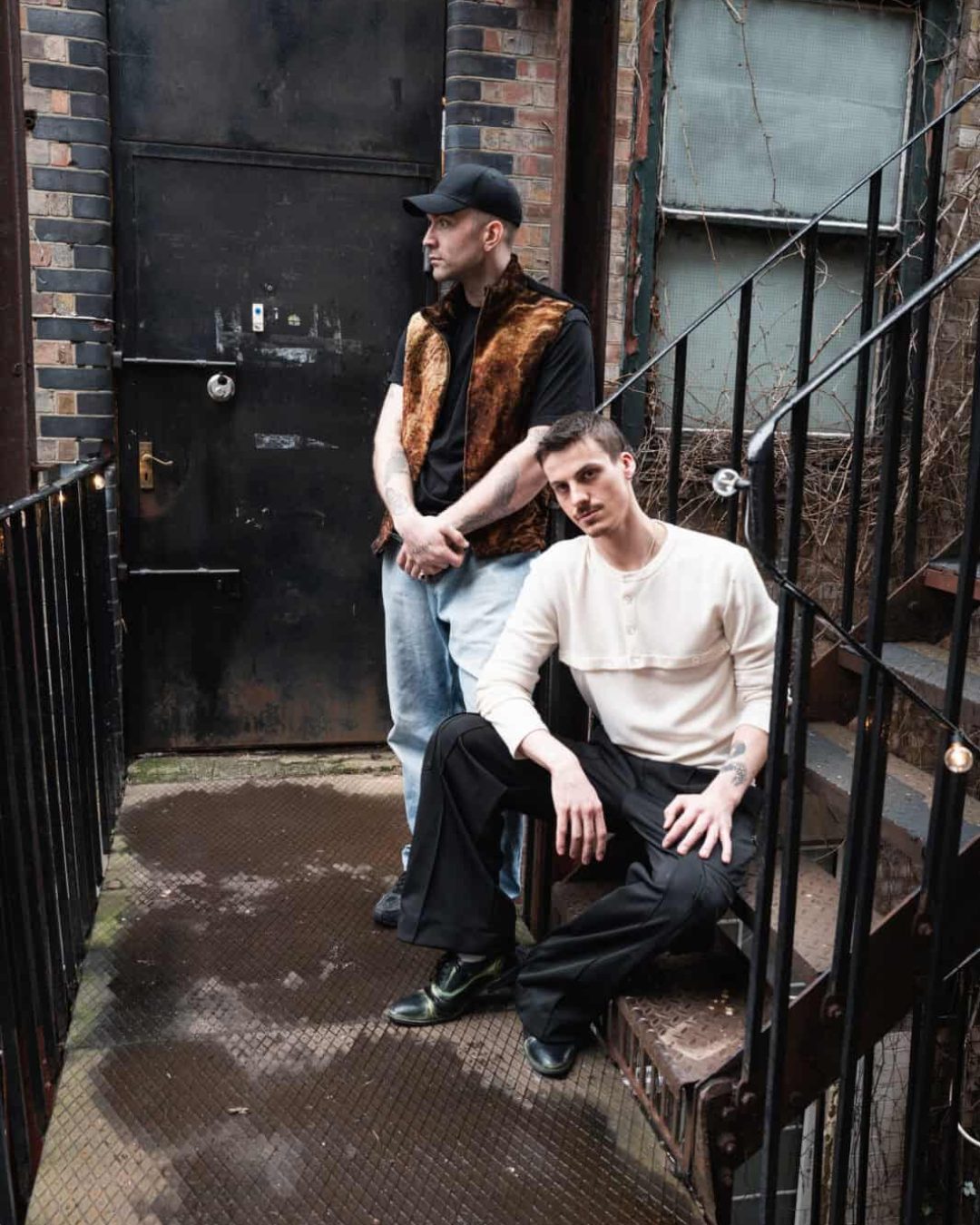 Two men pose on a metal stairway and landing outside a brick building. One stands by a black door, wearing a brown vest and cap, while the other sits on the steps in a white shirt and black pants.