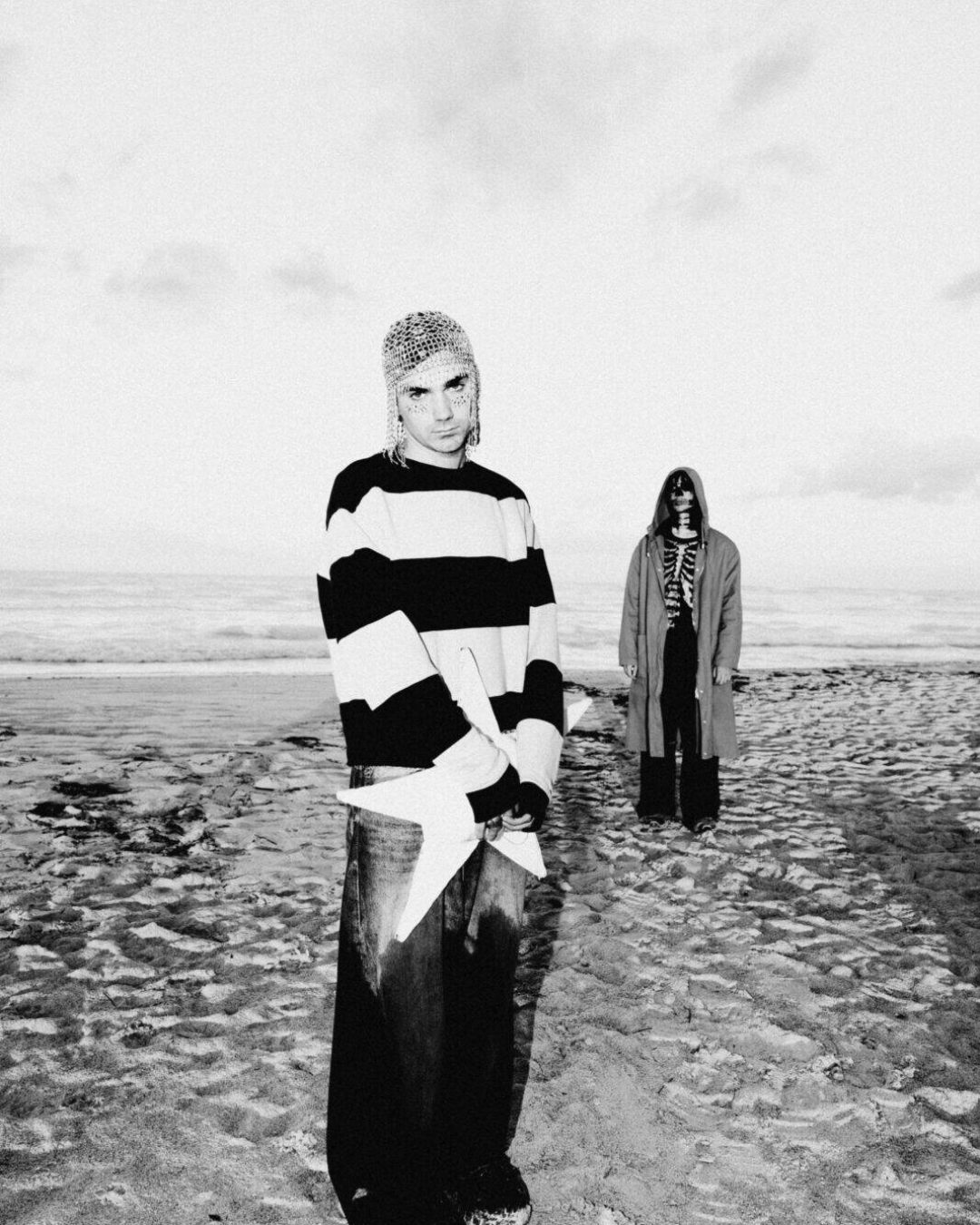 Piccolo and a second person stand on a beach in black-and-white. Piccolo wears a striped sweater and a chainmail-like headpiece. The second person, farther back, wears a long coat and sunglasses. The sky is cloudy.