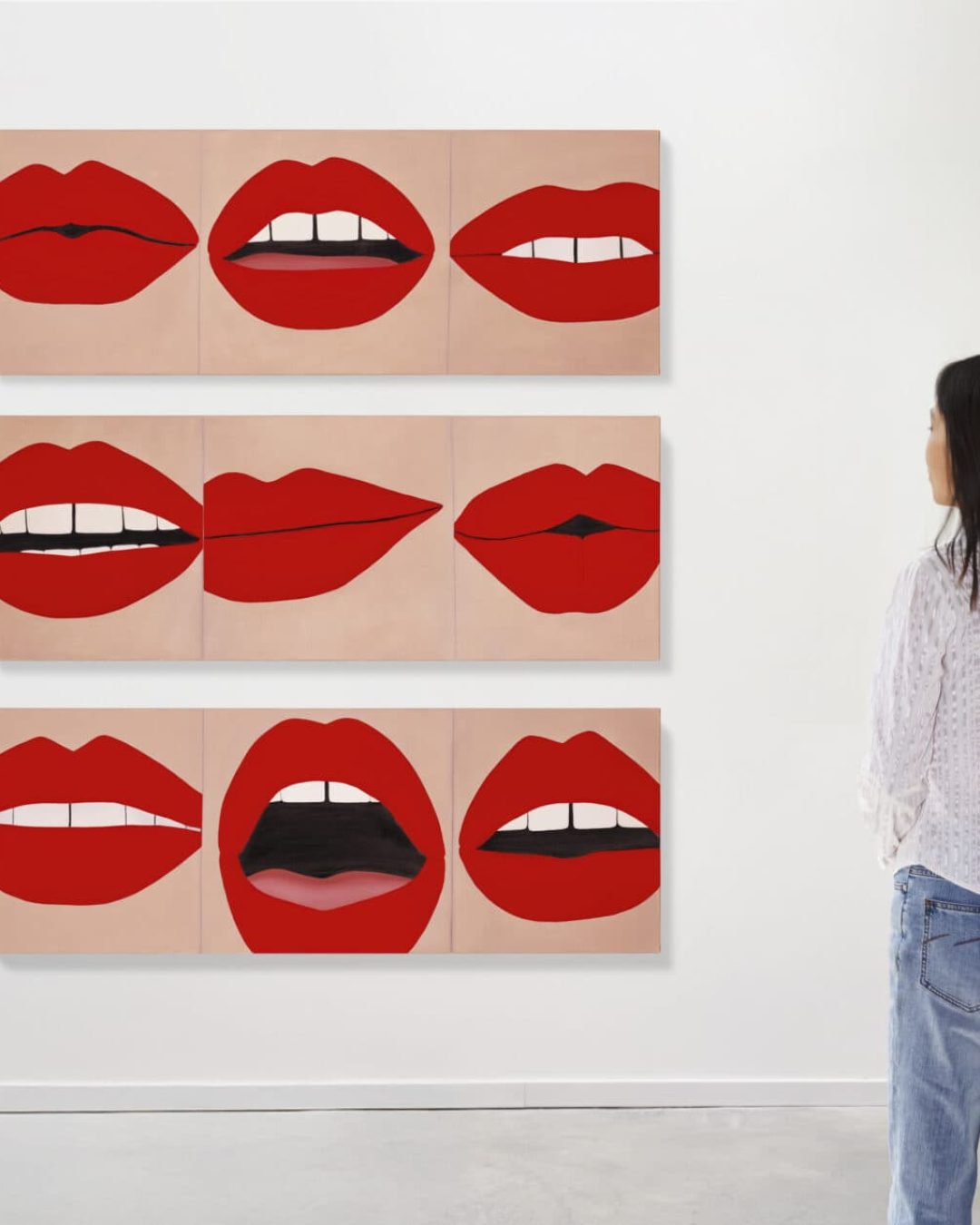 A woman stands in front of a wall displaying nine paintings of red lips in various shapes and expressions, arranged in three horizontal rows.