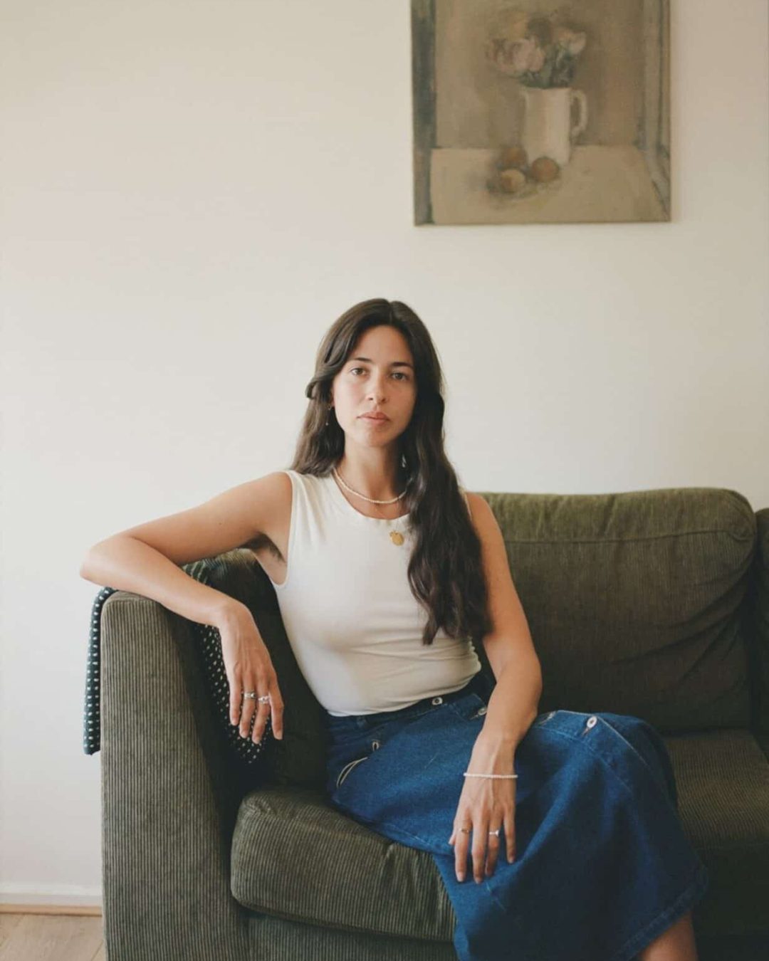 A woman with long dark hair sits on a green sofa, wearing a white sleeveless top and a blue skirt. She looks at the camera with a neutral expression. A painting of flowers in a vase hangs on the wall behind her.