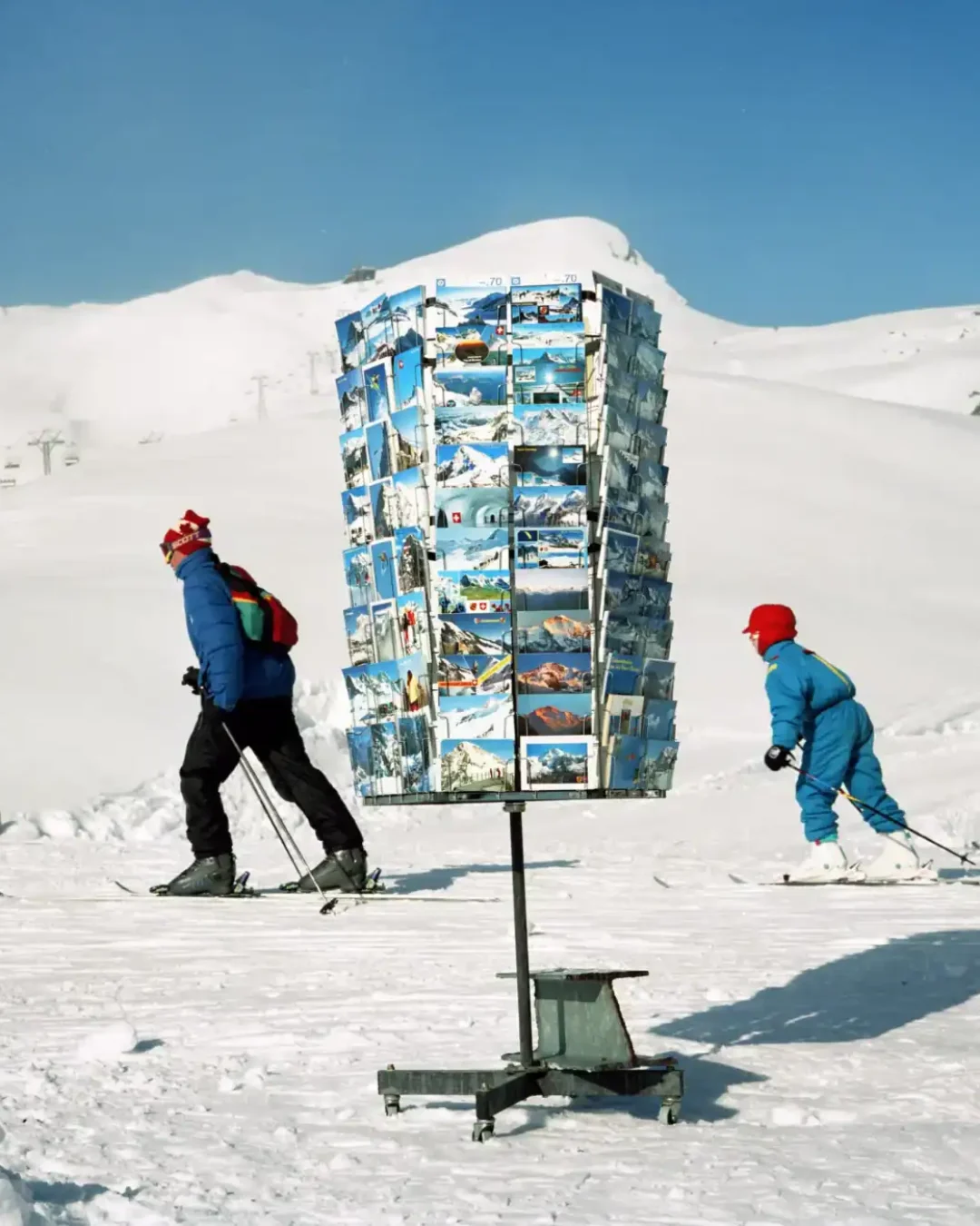A rotating stand filled with postcards stands on snowy ground while two people in colorful winter gear ski past it; snow-covered mountains rise in the background under a clear blue sky.