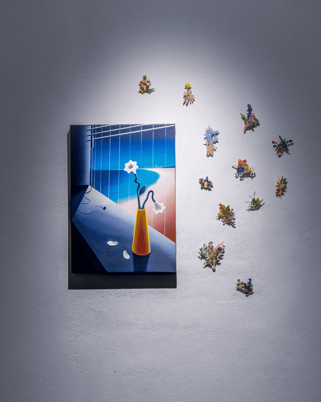 A painting by Hiroki Yagi of a yellow vase with white flowers sits on a dark table, hung on a light gray wall. Colorful, small, flower-shaped decorations are arranged on the wall to the right of the painting.