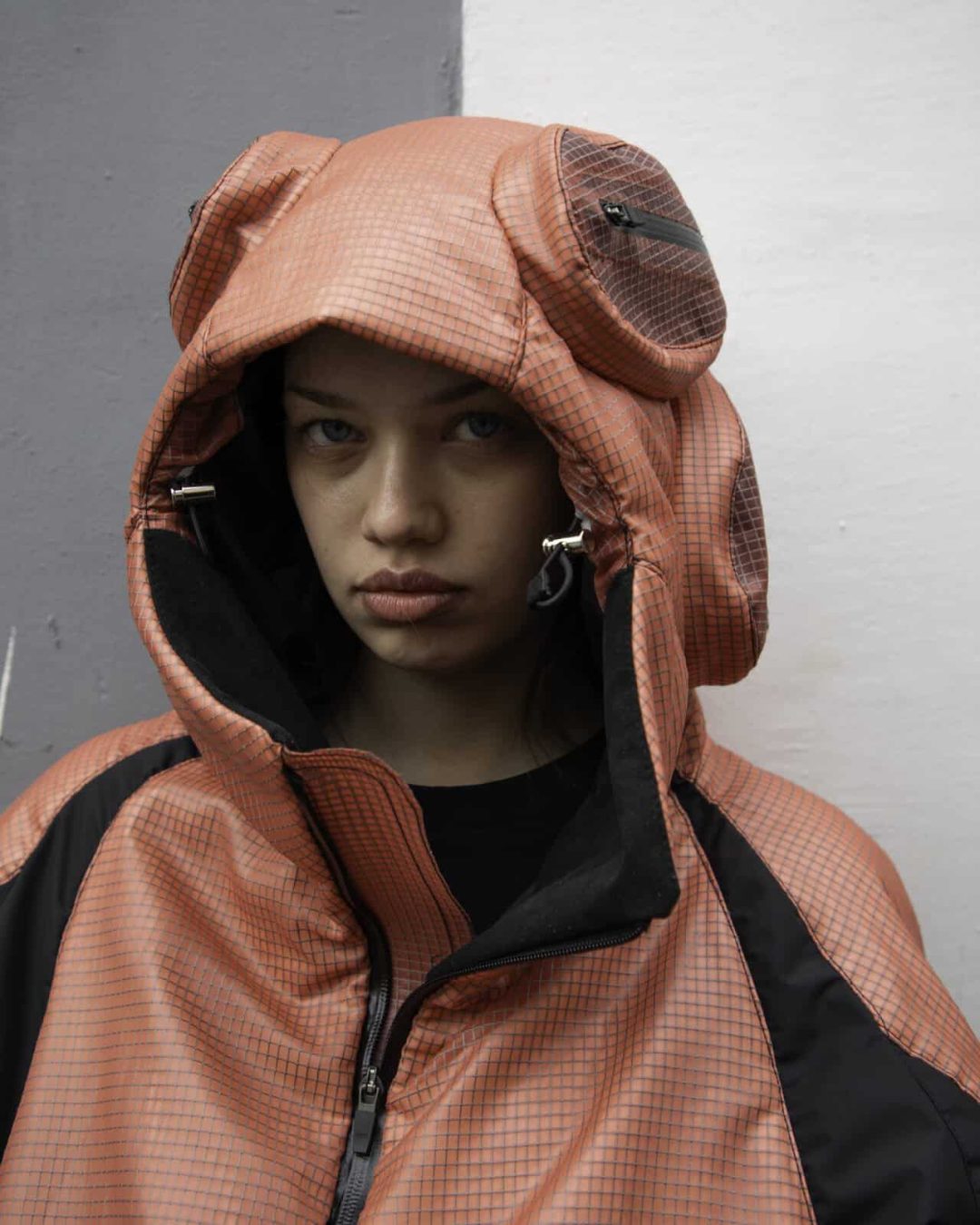 A person wearing a coral pink hooded jacket with large, circular ear-like shapes on the hood stands against a background divided into gray and white. The person looks directly at the camera with a serious expression.