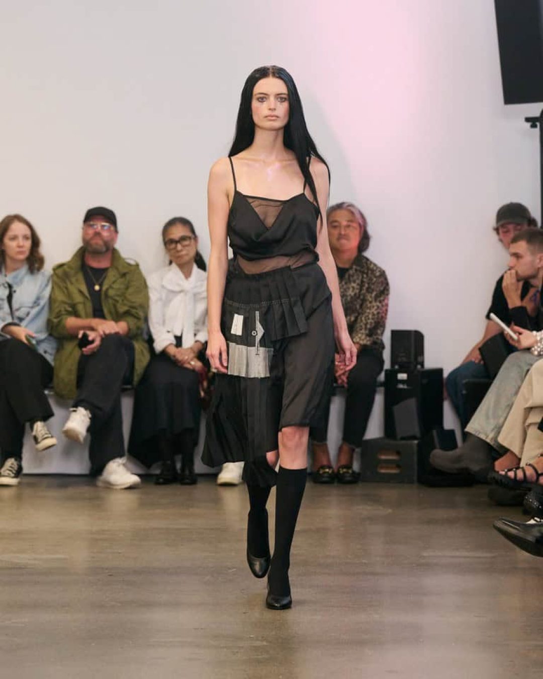 A model with long dark hair walks a runway wearing a sheer black dress with layered fabric details and knee-high black socks. Seated audience members watch from both sides of the runway in a minimalist room.