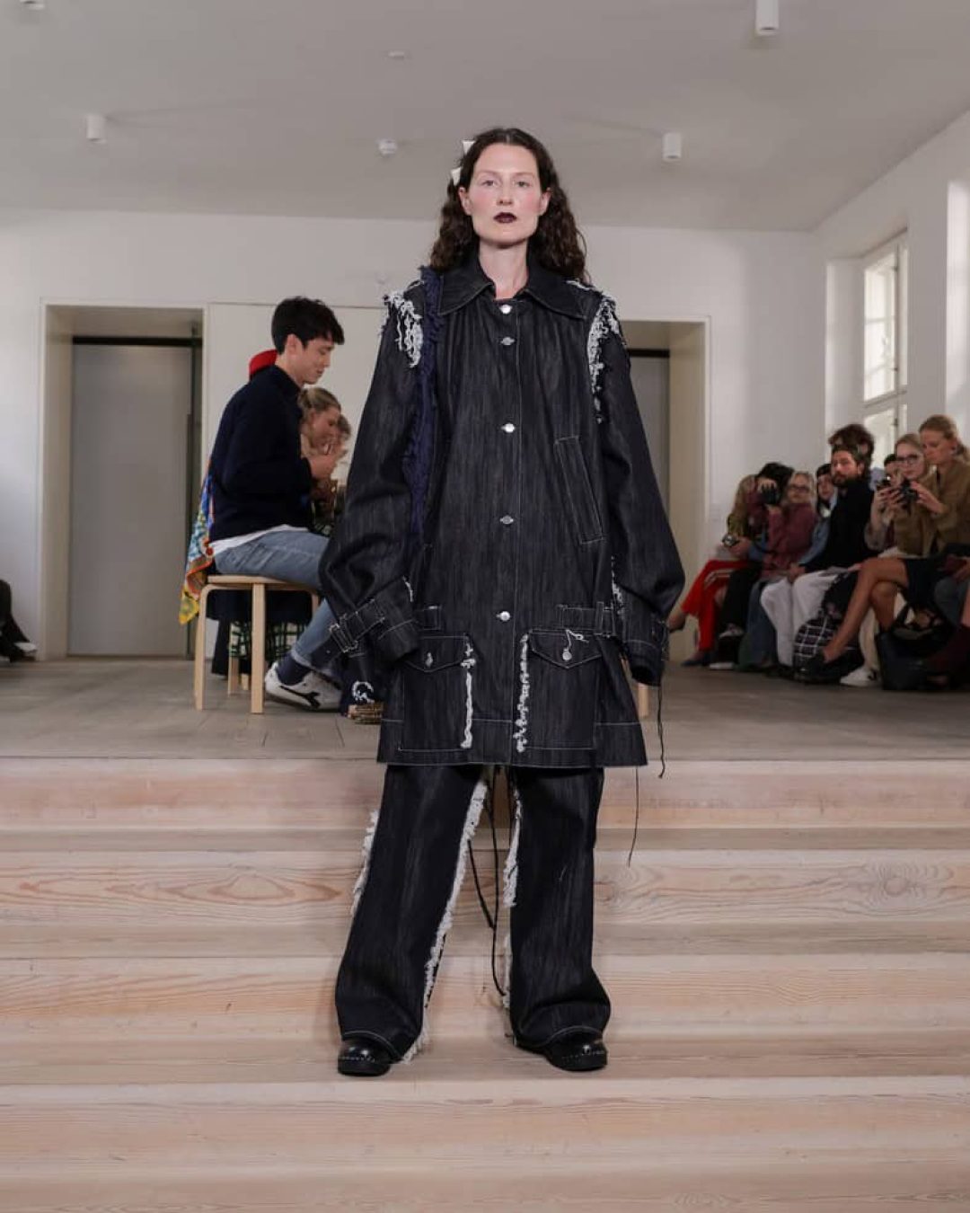 A model stands on wooden steps wearing an oversized black denim jacket and matching wide-legged pants with white stitching, in front of a seated audience at a fashion show in a minimalist indoor space.