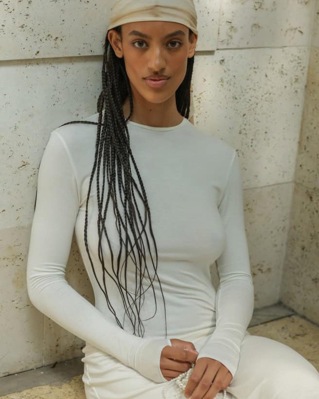 A woman with long braided hair, wearing a cream-colored headscarf and a fitted long-sleeve white dress, is seated against a textured stone wall, holding a beaded Taus necklace in her lap.