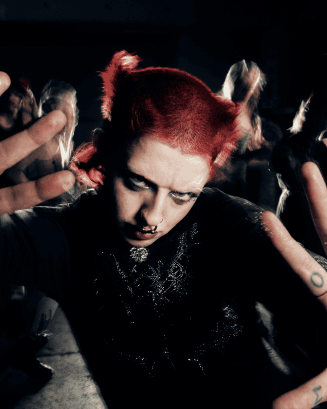 Cody Frost with bright red hair, dark eye makeup, facial piercings, and rings on their fingers poses with hands outstretched toward the camera at a crowded event, with people blurred in the background.