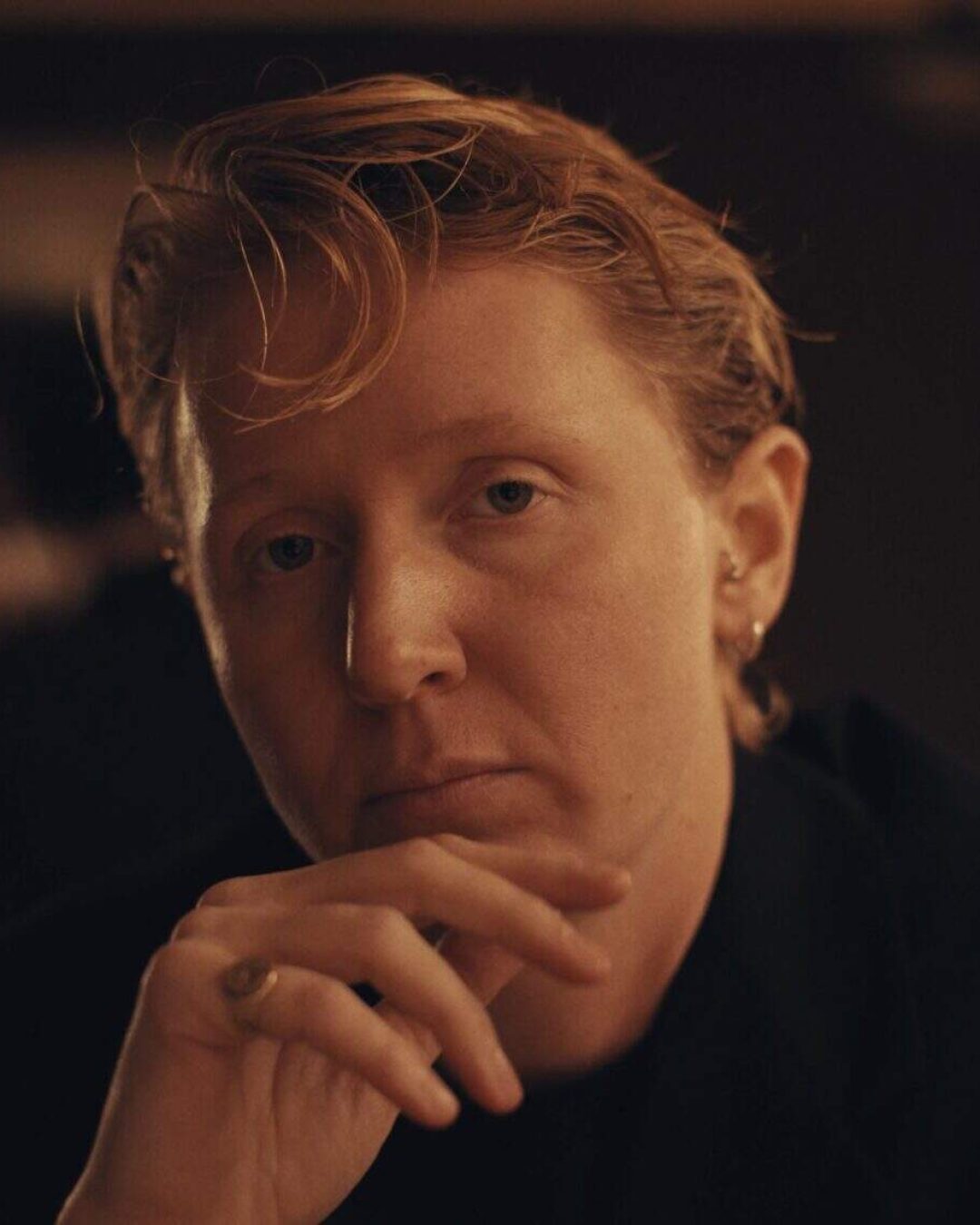 A person with short, wavy reddish-blonde hair and light skin looks thoughtfully at the camera, resting their hand near their chin in a dimly lit setting.