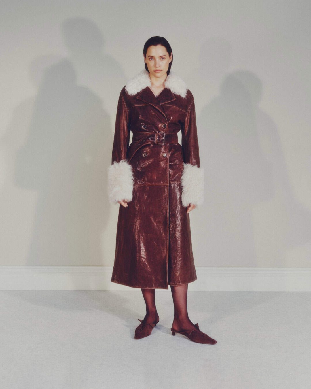 A woman stands against a light-colored wall, wearing a long brown leather coat with a wide belt, white faux fur collar and cuffs, black tights, and brown heeled shoes. Her expression is neutral.