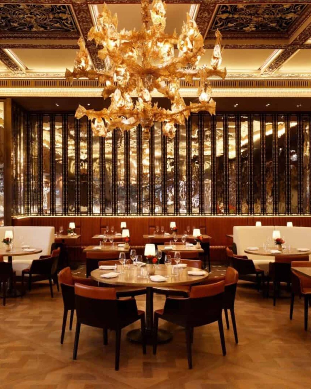 Elegant restaurant interior with round tables, white booths, brown chairs, a grand golden chandelier, warm lighting, wooden floors, and decorative gold accents. Red flowers and table settings add a refined touch, advice about where to eat during London Fashion week.