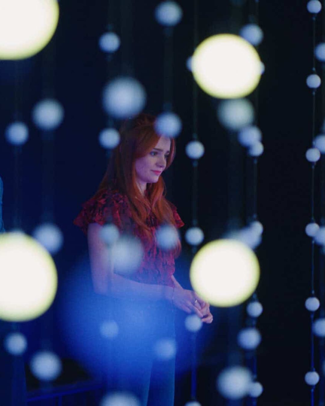 A man and a woman stand in a dimly lit room, partially obscured by hanging strings of glowing orbs. The background is dark, highlighting the illuminated orbs and creating a dreamy, contemplative atmosphere.
