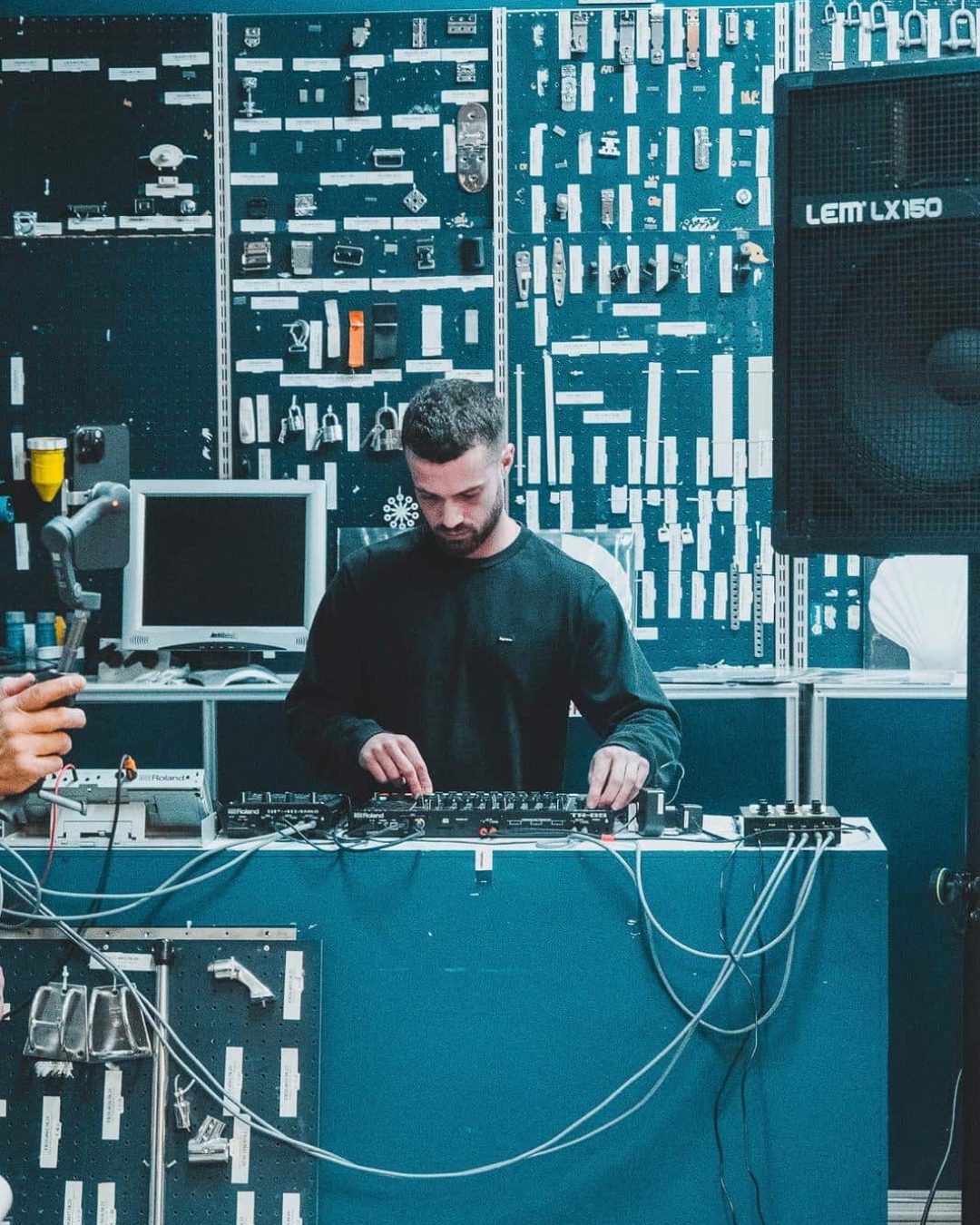 A man stands behind a DJ mixer, adjusting controls. He is surrounded by wires and electronic equipment, with a wall covered in tools and devices in the background. A large speaker is visible on the right.