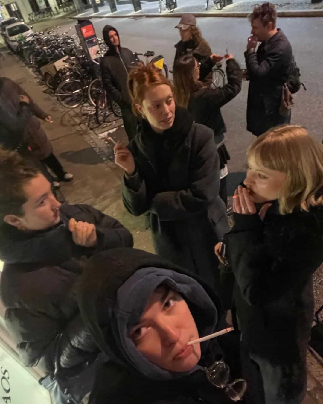 A group of young people stand outside at night in heavy coats. Some are smoking and talking, while bicycles are parked along the sidewalk behind them. The scene appears casual and social.