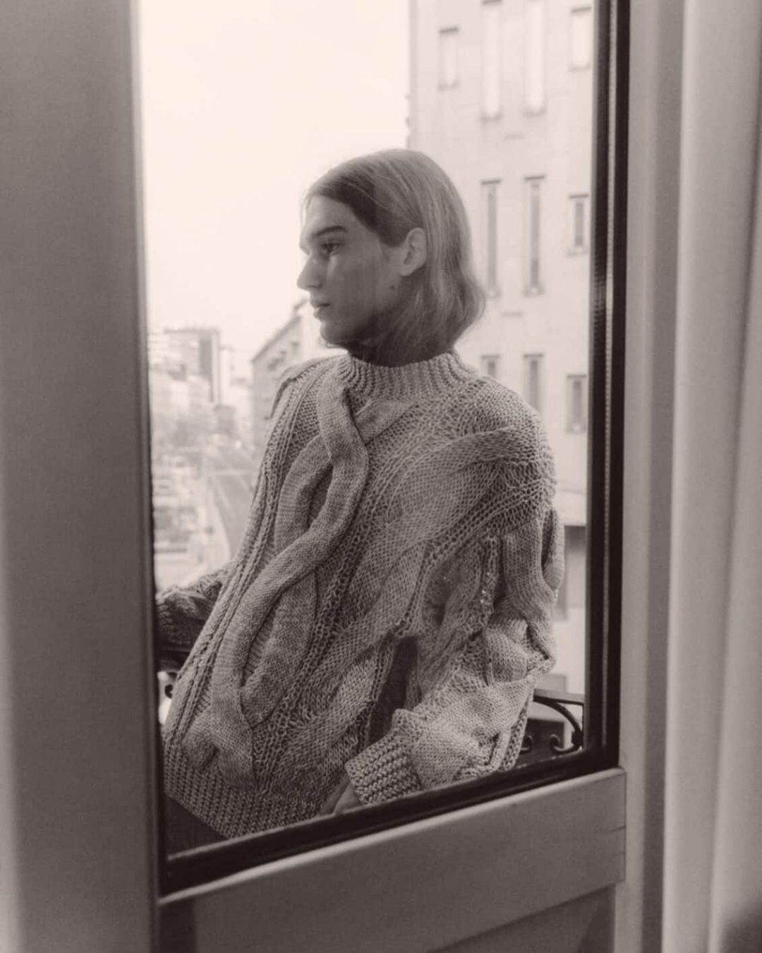 A person with long hair stands outside on a balcony, wearing a thick, cable-knit sweater, looking thoughtfully into the distance. The scene is viewed through a window, and the cityscape is visible in the background.