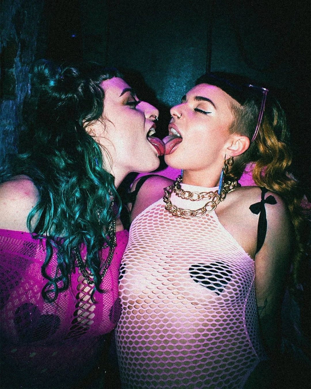Two women with vibrant hair and bold makeup stand close together at a party, dressed in mesh tops and accessories, playfully touching tongues in a colorful, nightclub-like setting.
