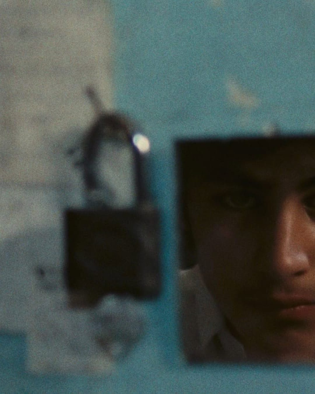 A young person’s face is partially reflected in a small, square mirror on a blue wall with faded papers and a black padlock hanging nearby. The expression appears serious and contemplative.