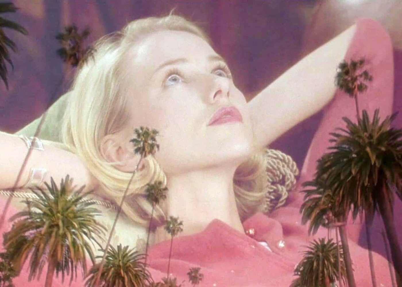 A blonde woman in a pink top reclines with her hands behind her head, gazing upward dreamily. Semi-transparent palm trees overlay the image, giving it a surreal effect reminiscent of a still from Mulholland Drive.