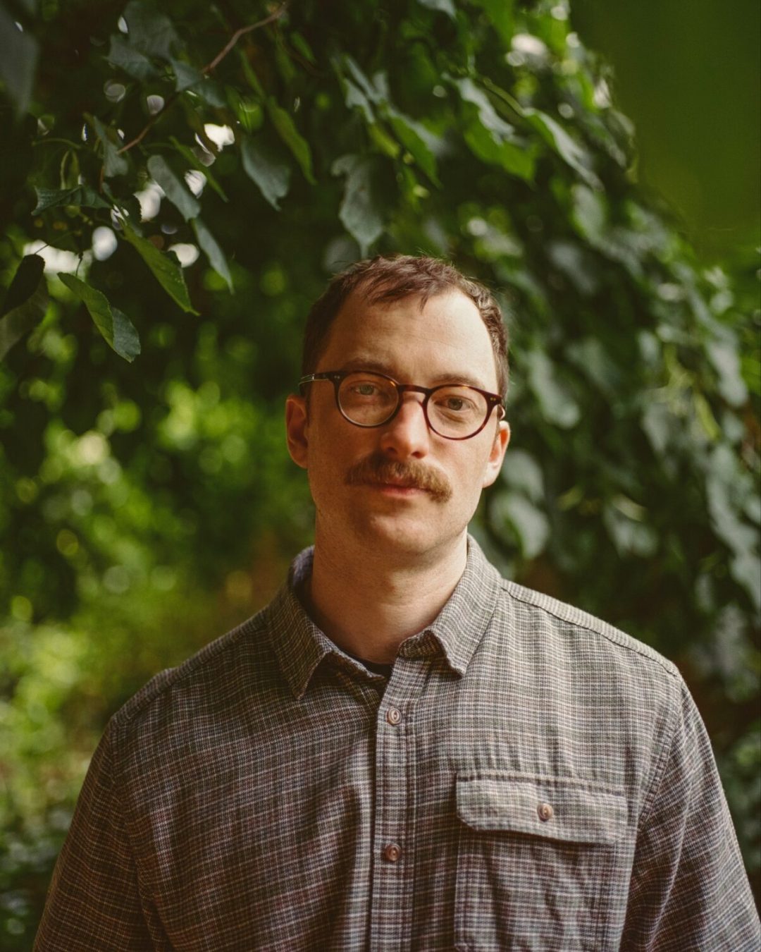 Author Jem Calder posing for a portrait in a gray plaid shirt and glasses against a lush green foliage background.