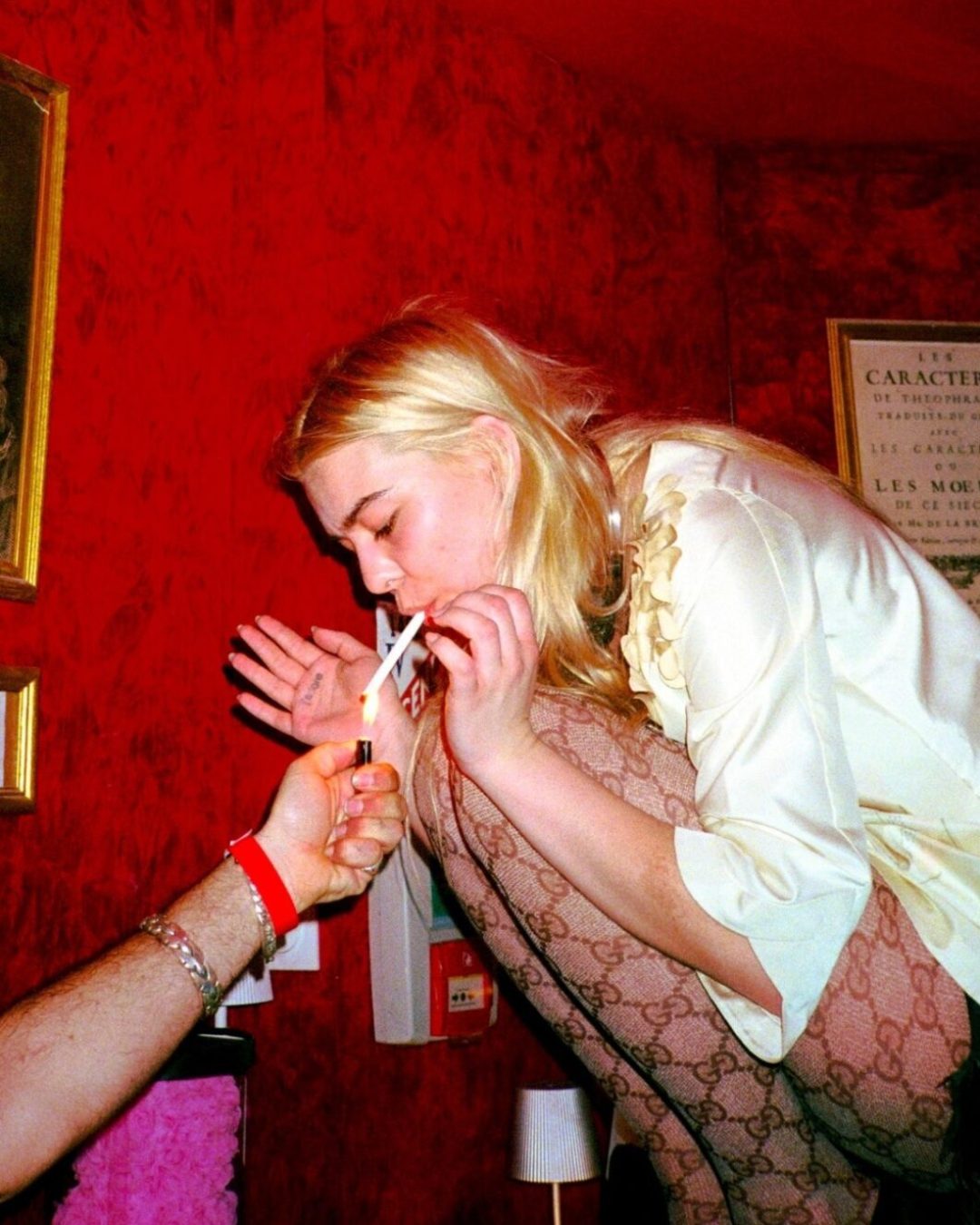 A blonde woman in a ruffled cream blouse and Gucci monogram tights leaning forward to light a cigarette from a hand extended toward her, against a deep red wall — photograph by Arthur Samier
