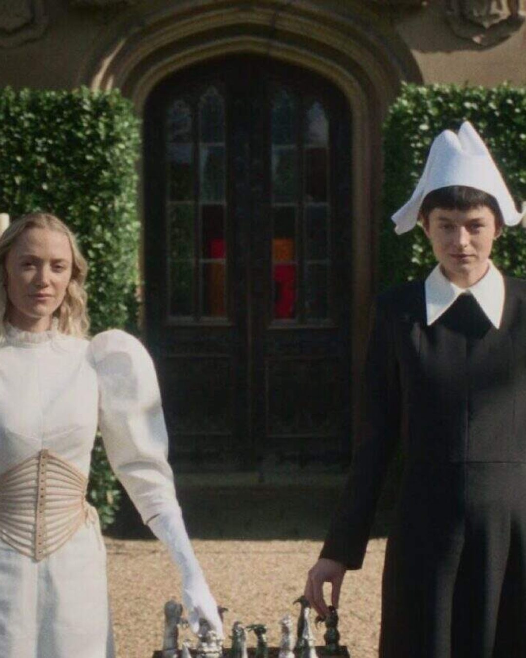 Two women stand side by side outdoors: one in a white dress with puffed sleeves and gloves, the other in a black dress and white bonnet, holding a bird of prey. A chessboard sits in front of them, with a mansion and greenery behind.