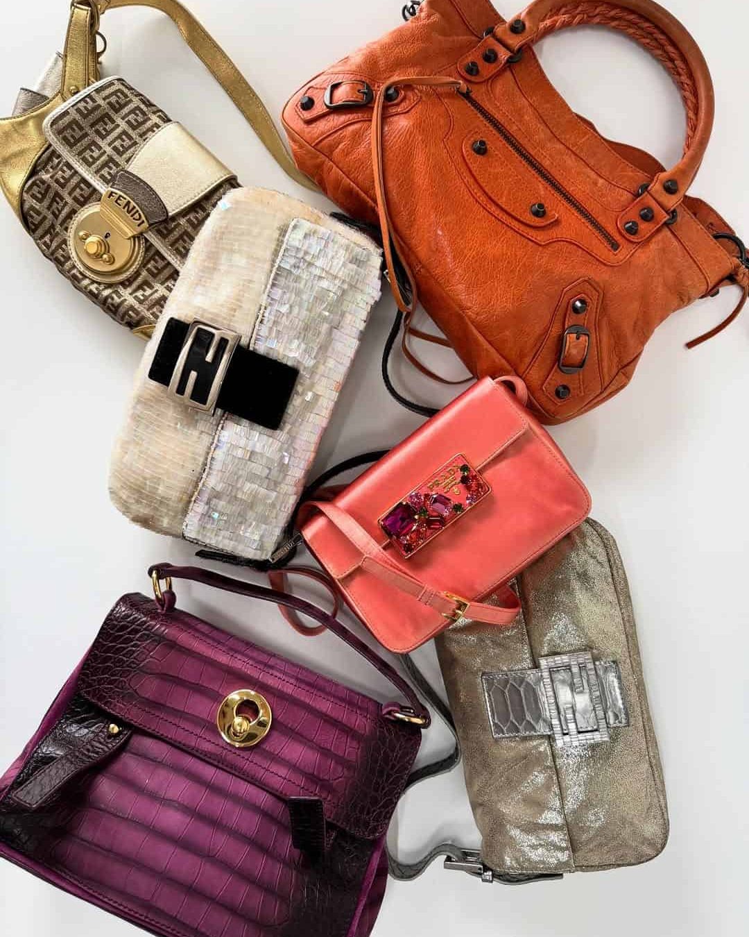 Six designer handbags in various colors and styles are arranged on a white surface, including metallic gold, woven white, coral, orange, deep purple, and silver bags with distinctive clasps and hardware.