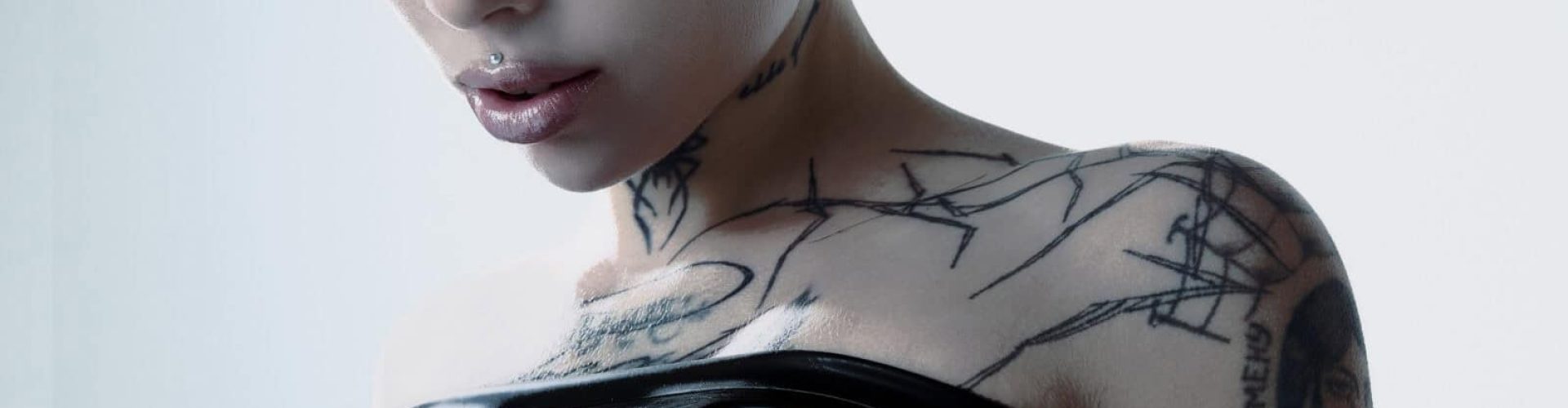 A close-up of a person with short hair, a nose piercing, and tattoos on their neck and shoulder. They are wearing a shiny black off-shoulder top, and water droplets are visible on their skin.