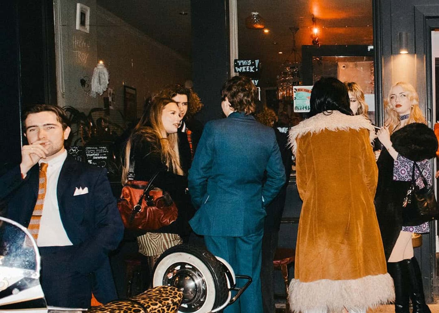 A stylish group of people gathers outside a dimly lit café at night; one Tickety Boo man in a suit stands near a white scooter with a leopard print seat, while others chat by the entrance, dressed in fashionable retro clothing.